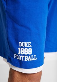 Pantalones cortos deportivos azules con el texto blanco "DUKE 1888 FOOTBALL" llevados sobre pantalones cortos de malla blancos en la pierna de una persona.
