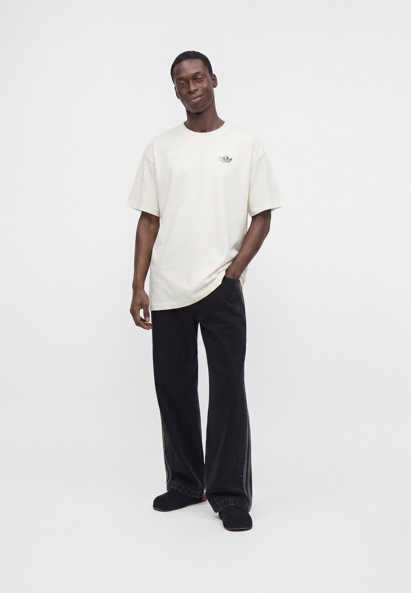 Uomo in piedi con una mano in tasca che indossa una T-shirt oversize color avorio e pantaloni neri larghi su sfondo bianco.