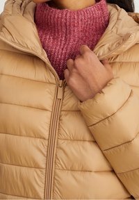 Chaqueta acolchada beige con costuras horizontales y cierre con cremallera, superpuesta sobre un suéter de cuello alto rosa texturizado.