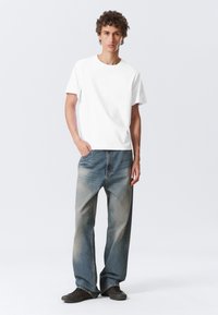 Weißes Baumwoll-T-Shirt mit Rundhalsausschnitt, kombiniert mit verwaschenen blauen Jeans und dunklen Schuhen. Das Model steht mit einer Hand in der Tasche.