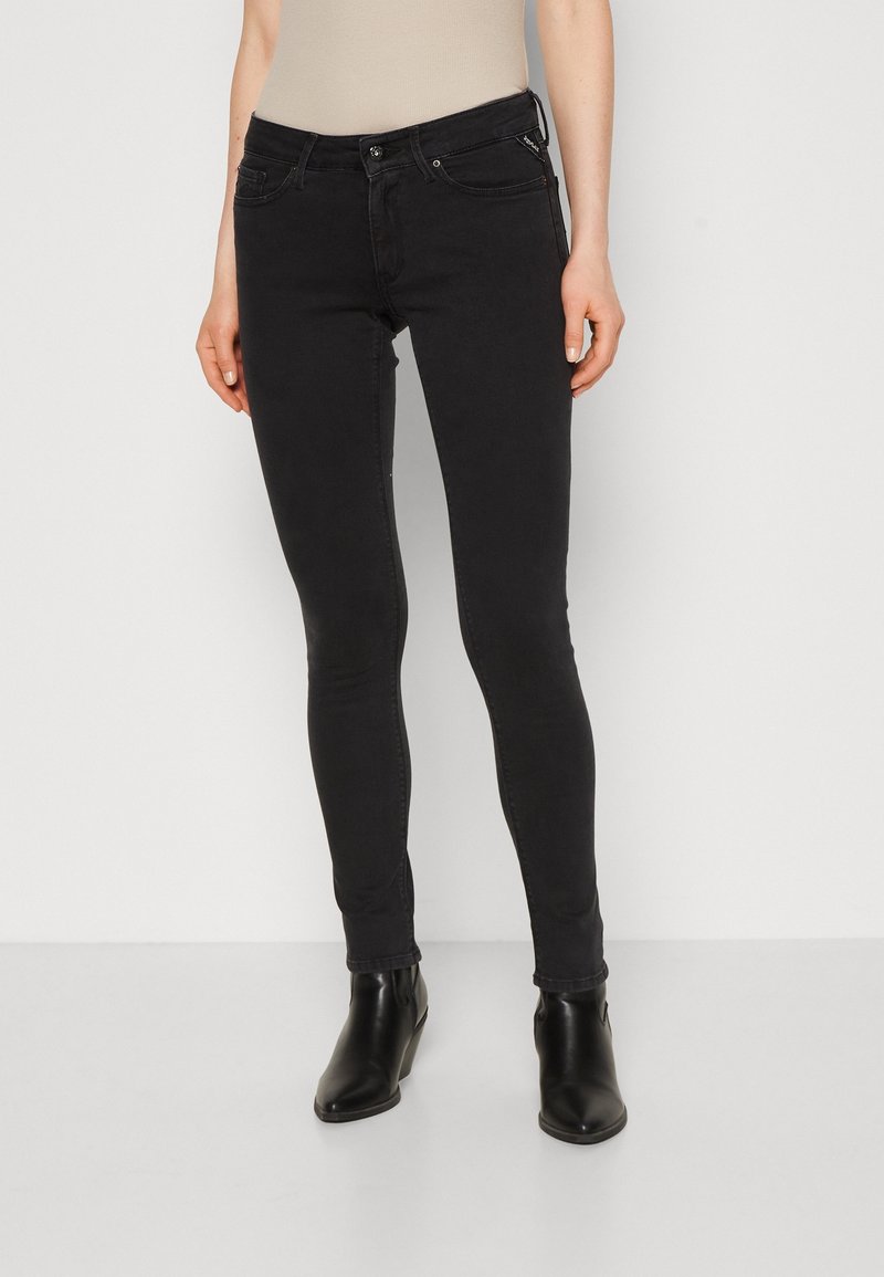 Femme portant un jean noir ajusté et des bottines noires, debout devant un fond uni clair.