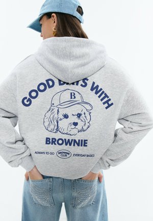 Persona con una sudadera gris con capucha que tiene una ilustración de un perro y el texto "Buenos Días con Brownie" en la espalda, combinada con jeans de mezclilla con tachuelas.