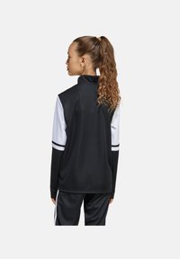Veste de sport noire avec des accents de manches blancs et noirs, col haut et poignets élastiques. Le design présente trois bandes sur chaque manche.