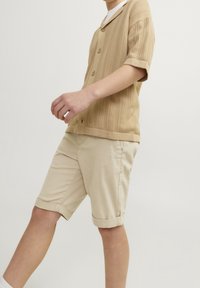 Beige gebreid shirt met korte mouwen en een kraag, featuring een textuurpatroon en knoopsluitingen. Beige shorts met opgerolde mouwen complementeren het bovenstuk.