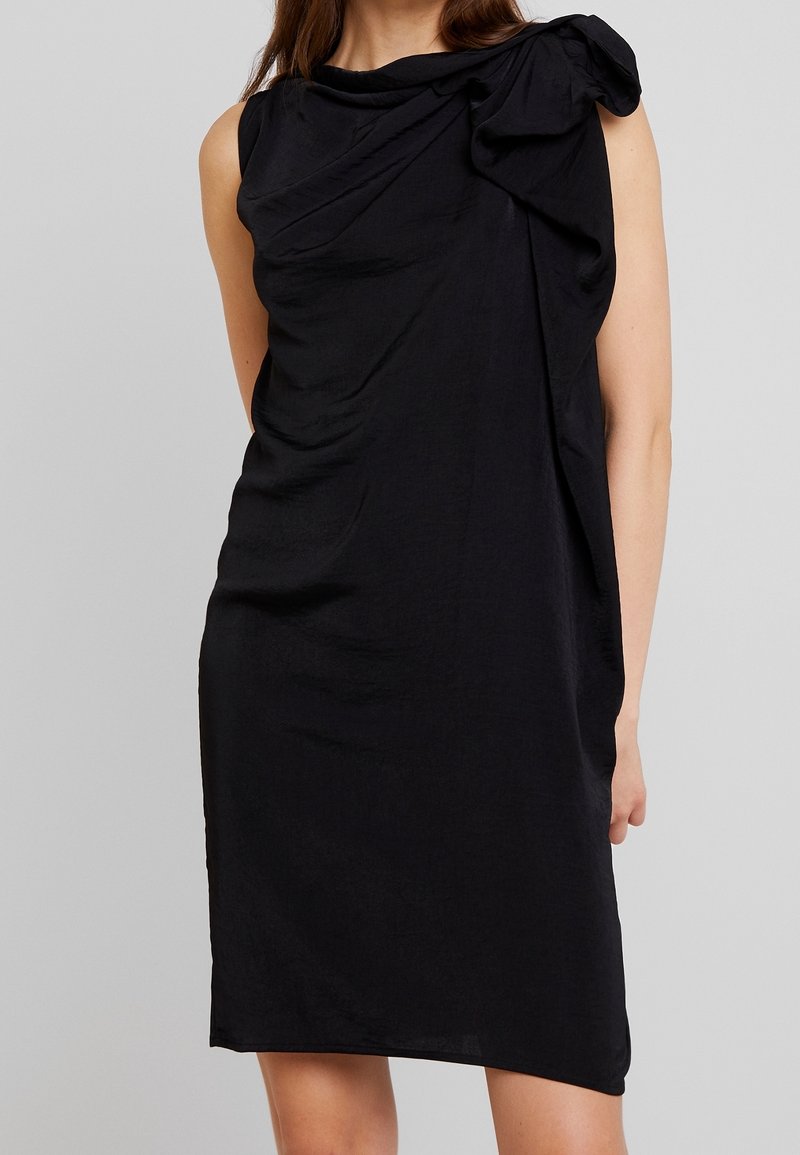 Robe noire sans manches avec un design drapé et un détail noué sur une épaule, fabriquée à partir d'un tissu doux et léger. Texture lisse, longueur au genou.