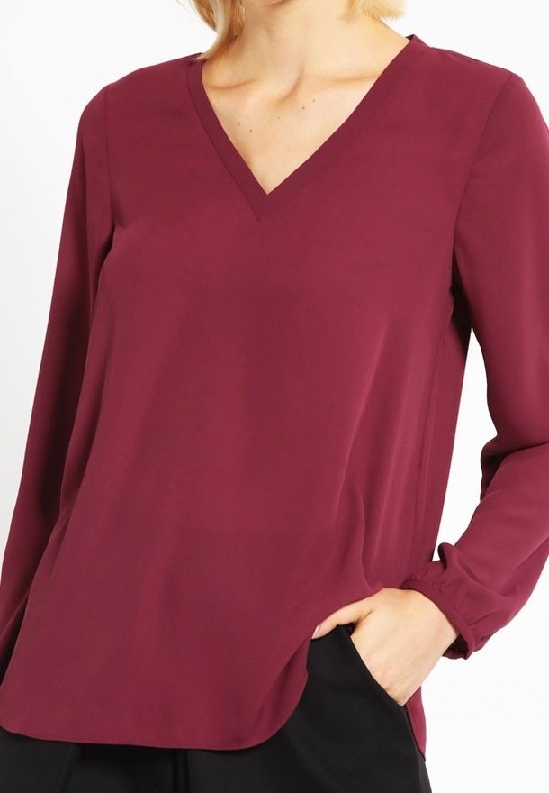 Blouse bordeaux à col en V avec manches longues, en tissu lisse. Présente une coupe décontractée et un léger éclat, avec un ourlet droit et des fentes latérales.
