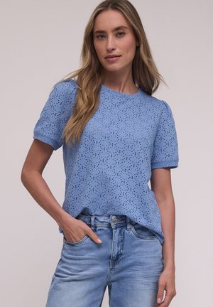 Kvinde med langt blondt hår iført en lys blå perforeret top med korte ærmer og lyseblå denimjeans, stående med den ene hånd i lommen.