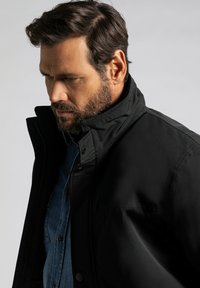 Schwarze Außensjacke mit hohem Kragen, ausgestattet mit einem Knopfverschluss und einer glatten Textur, über einem blauen Jeanshemd getragen.