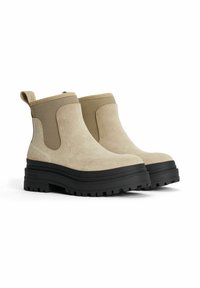 Beige ruskindsankelevarer med sort gummisål, kraftig sål, elastiske sidepaneler og en løkke ved hælen. Glat tekstur med mat finish.