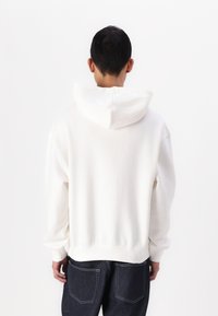 Sweatshirt à capuche blanc avec une coupe décontractée, texture douce et poignets côtelés. L'arrière présente un design simple, associé à un pantalon en denim noir.