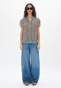 Blusa a fantasia nera e crema con maniche a ruches, indossata con jeans a gamba larga blu e sandali neri. Sfondo bianco, senza accessori.