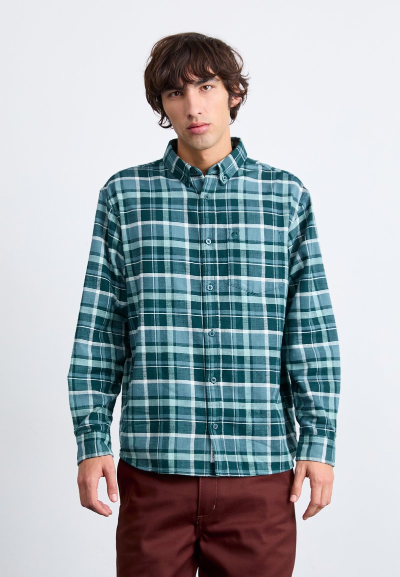 Chemise à carreaux verte et bleue à boutons, à manches longues, avec un col et une poche sur la poitrine, fabriquée dans un tissu doux.