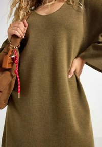 Oliven grøn oversized striktrøje i blødt strikket materiale med scoop-neck og sidelommer. En brun ruskinds taske med en rød accentrem.