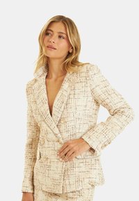 Blazer en tweed beige clair avec une coupe structurée, design à double boutonnage, tissu texturé et boutons dorés.