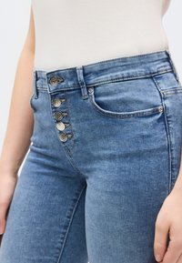 Magas derekú kék farmernadrág, öt gombos sliccel, övbújtatókkal és elülső zsebekkel. A szövet sima textúrájú, enyhén kifakult.