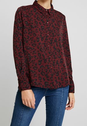 Frau trägt ein bordeauxrotes, langärmliges Hemd mit schwarzem Leopardenmuster, kombiniert mit blauen Jeans, und steht vor einem schlichten grauen Hintergrund.