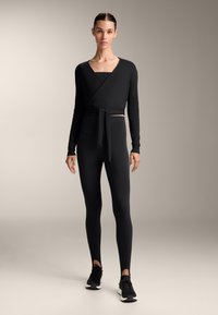 Fekete, hosszú ujjú, megkötős felső, derékon megkötéssel, fekete, magas derekú leggings-szel párosítva, mindkettő rugalmas anyagból készült, és fekete sportcipővel.