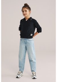 Noor tüdruk seisab käed selja taga, seljas must kraega kampsun, seljas hele sinised teksad ja jalas hall-valged tossud ühtlase taustaga.