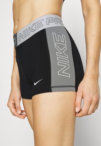 Femme portant un short de sport Nike Pro noir et gris avec des logos sur la ceinture, le panneau latéral gauche et la partie inférieure avant de la jambe.