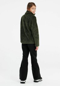 Grüne Fleecejacke mit hohem Kragen und strukturiertem Finish, kombiniert mit schwarzen Hosen und Stiefeln mit weißer Sohle. Modell von hinten gezeigt.