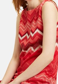 Czerwona sukienka bez rękawów z wzorem w zygzaki w różnych odcieniach czerwieni i bieli. Materiał jest teksturowany i lekki.