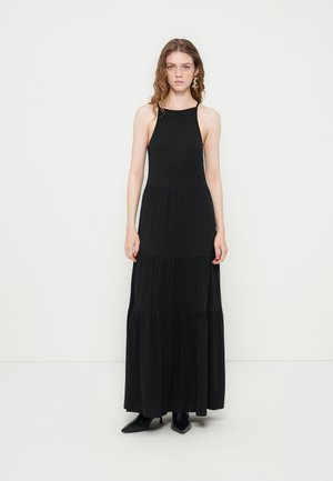 Jeune femme aux cheveux ondulés portant une robe longue noire sans manches à volants et des bottes noires pointues, debout devant un fond clair uni.