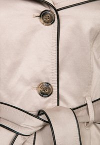 Beżowy płaszcz trencz z czarnym obszyciem, z okrągłymi metalicznymi guzikami z logo. Zawiera dopasowany pasek i wytrzymałą teksturę tkaniny.