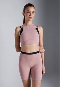 Svetlo roza rebrast športni crop top z visokim ovratnikom in črnimi naramnicami, kombiniran z ustreznimi visokim pasom kratkimi hlačami s črnimi detajli.