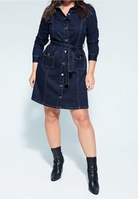 Robe chemise en denim avec des manches longues, fermeture par boutons, deux poches avant et une ceinture en tissu. Couleur bleu foncé avec des surpiqûres visibles.