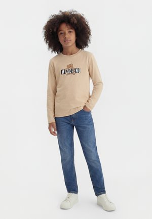 Barn med krøllet hår iført beige langærmet trøje med bamseprint og blå jeans, stående med den ene hånd i lommen på hvid baggrund.