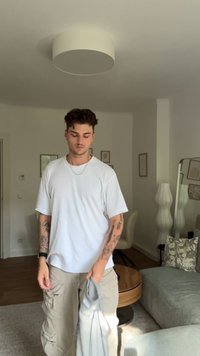 Bílé tričko s krátkým rukávem, béžové cargo kalhoty se zipy, světlá pleť, vlasy střední délky a kudrnaté, v jasné místnosti s minimálním dekorem.