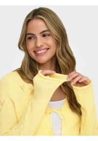 Yellow knitted cardigan with a textured finish, featuring long sleeves and a tie closure at the neckline. Light layered top underneath.