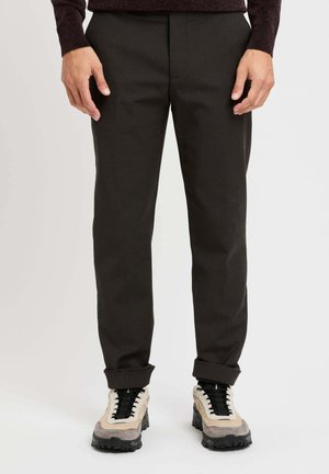 Pantaloni marroni scuri con una vestibilità slim, caratterizzati da una texture liscia e pieghe frontali. Abbinati a scarpe sportive beige e grigie con lacci neri.