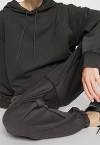 Persona che indossa un felpa grigia scura con un logo floreale e pantaloni jogging coordinati, seduta con una gamba incrociata e la mano appoggiata sul ginocchio.