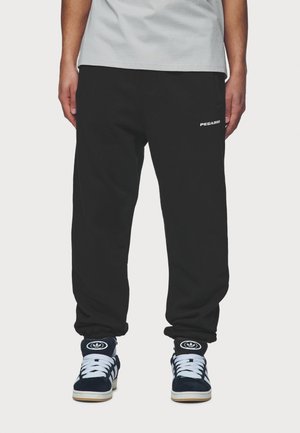 Mężczyzna w czarnych spodniach dresowych Pegador i granatowych butach sportowych Adidas, stojący na jednolitym jasnym tle.