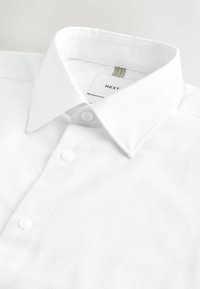 Fehér gombos ing hegyes gallérral, két látható gombbal és a gallér belsejében látható "NEXT" márkajelzéssel.