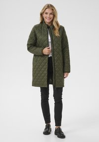 Manteau matelassé vert olive avec un col haut, une fermeture éclair et des poches latérales. Porté sur un haut blanc et un pantalon noir, avec des chaussures noires brillantes.