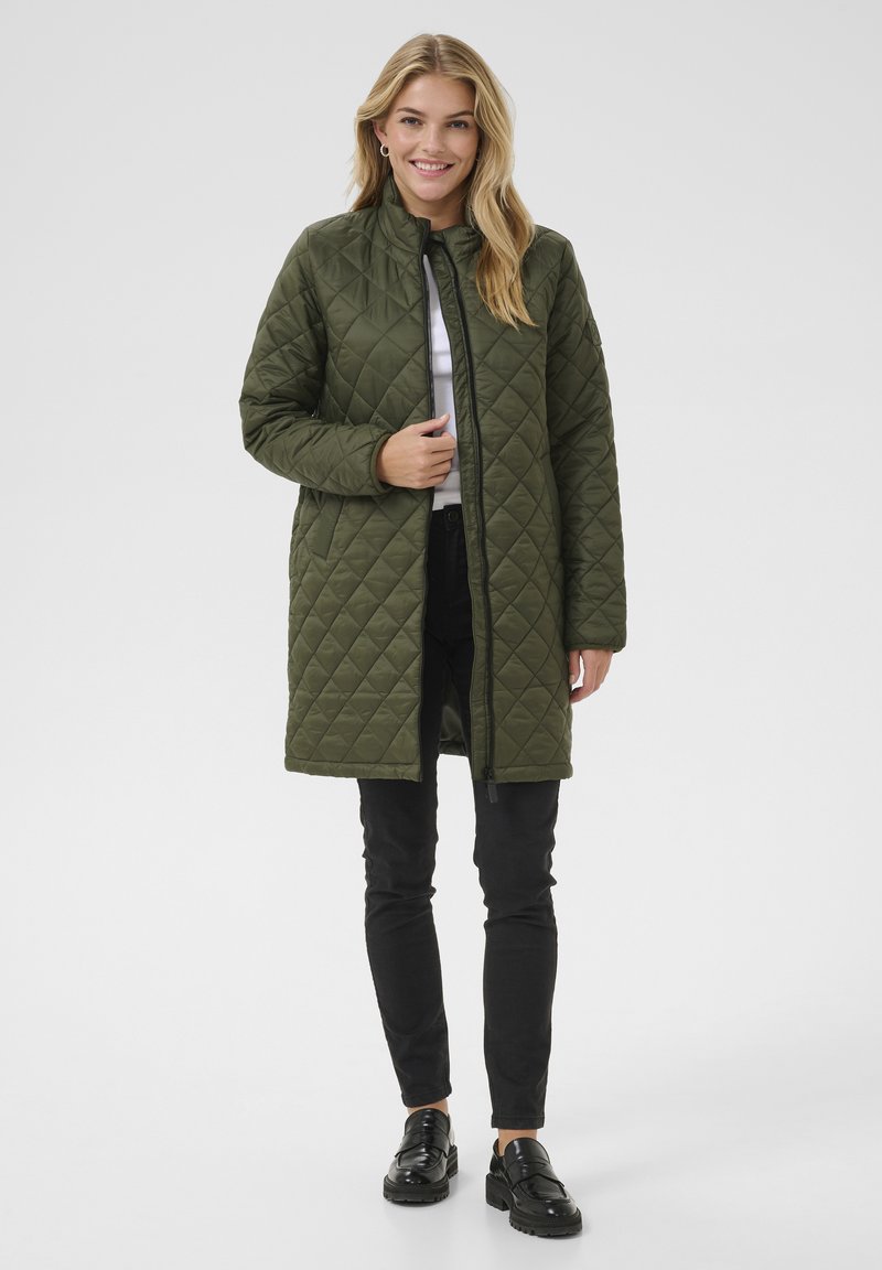 Manteau matelassé vert olive avec un col haut, une fermeture éclair et des poches latérales. Porté sur un haut blanc et un pantalon noir, avec des chaussures noires brillantes.