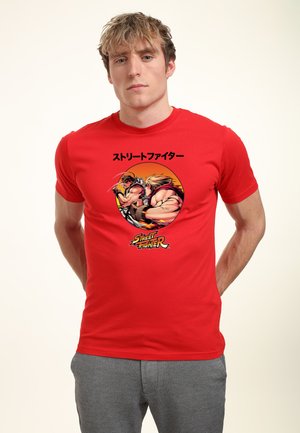 Junger Mann trägt rotes T-Shirt mit einem runden Street Fighter-Design, das zwei kämpfende Kämpfer zeigt, mit japanischem Text darüber.