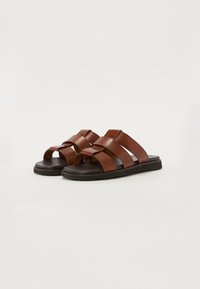 Sandales en cuir brun avec des brides croisées, design ouvert et semelle texturée noire. Présentent un look simple et moderne avec une construction plate.