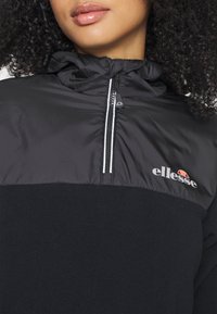 Gros plan d'une femme portant un pull polaire noir ellesse avec une demi-fermeture éclair et un logo sur la poitrine, cheveux bouclés encadrant le visage.
