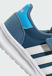 Detailný záber na modrú tenisku s bielou podrážkou a bielymi pruhmi, ktorá má polstrovaný golier a značkový modrý kožený pätový štítok.