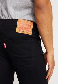 Jeans de mezclilla negra con una etiqueta de marca de cuero marrón, que incluyen bolsillos curvados y una etiqueta roja en el bolsillo trasero. La mano del modelo descansa en la cadera.