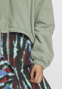 Sudadera verde recortada con cierre de cremallera, destacada por una manga texturizada. Combinada con una falda de patrón tie-dye colorida en negro, rojo y azul.