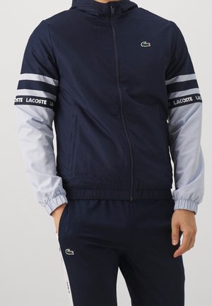 Muž nosící námořnicky modrou a světle šedou bundu na zip a kalhoty s logem Lacoste na hrudi, rukávech a straně kalhot.