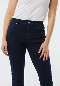 Jeans en denim bleu foncé avec un design ajusté, style à cinq poches et fermeture à bouton à la taille. Texture lisse et coupe ajustée.