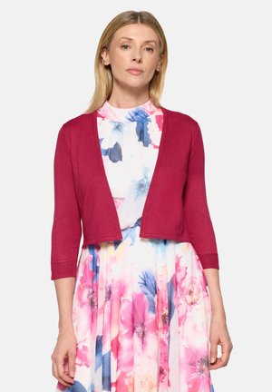 Kobieta w czerwonym krótkim kardiganie na kwiecistej sukience z wysokim kołnierzem, zdobionej dużymi różowymi, niebieskimi i białymi kwiatami, stojąca na jednolitym tle.