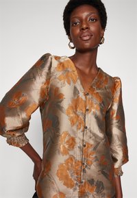 Blouse florale marron et dorée avec un décolleté en V, des manches bouffantes et des fermetures à boutons. Tissu texturé avec des motifs floraux complexes.