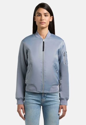 Kvinde med langt mørkt hår iført en lyseblå bomberjakke med lynlåslommer, ribstrikkede manchetter og blå jeans, stående mod en ensfarvet baggrund.