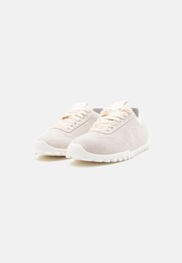 Paire de baskets en daim beige clair avec semelles blanches et lacets assortis sur un fond blanc uni, légèrement inclinées vers la droite.